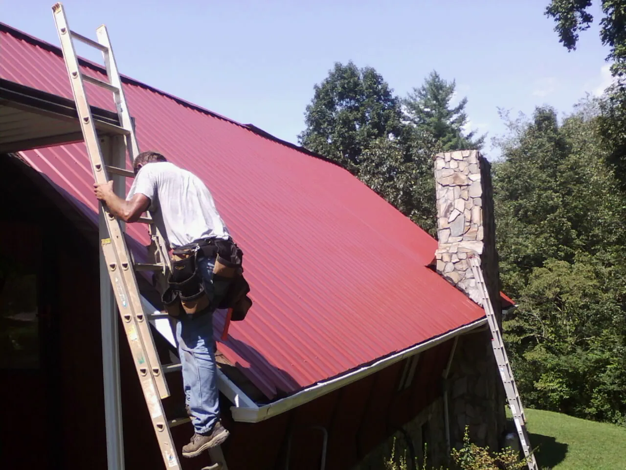 Roofer on a ladder beside a red metal roof and stone chimney — Soffit & Fascia Repair by Roofing Brothers in Dushore